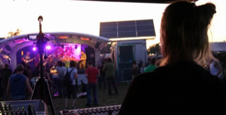 A stage lighting engineer stands at a lighting console, facing a stage with colorful lights and a crowd in the background during an outdoor festival event.